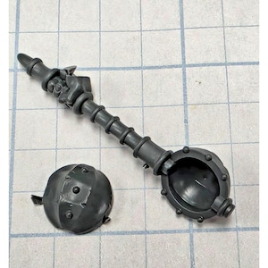 May include: A dark gray plastic miniature component featuring a long, segmented handle ending in a clenched fist, paired with a separate, hollow, spherical piece with small studs along its outer edge, designed for assembly in tabletop gaming.