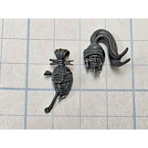 May include: Two unpainted gray plastic miniature heads. The left head features a topknot, a headband with a symbol, and a pipe held in the mouth. The right head features a wrapped head covering and a long, flowing ponytail extending to the right.