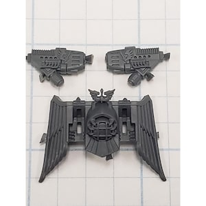 May include: A set of gray plastic miniature wargaming components. The set includes two detailed handguns and a winged emblem, likely for a tabletop game or model kit. The pieces are designed with intricate details.