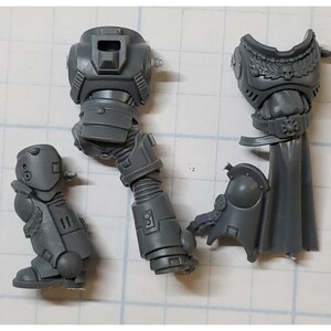 May include: A collection of unassembled gray plastic miniature figurines, including a leg, torso, and shoulder armor. The pieces are detailed with mechanical and decorative elements, suggesting a science fiction or fantasy theme.