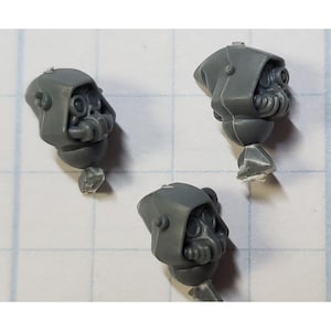 May include: Three gray plastic miniature heads with hooded helmets and gas masks. The helmets have a textured surface and the gas masks have a detailed design.
