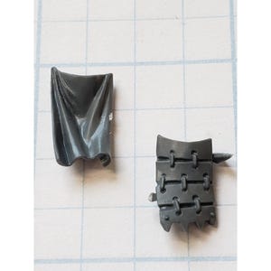 May include: Two small, gray plastic model pieces. One is a draped, rectangular shape, and the other is a segmented, rectangular piece with a series of linked loops and a small spike extending from the side. The pieces are on a grid background.