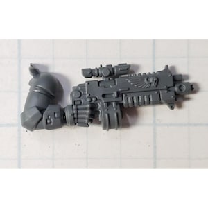 May include: A gray plastic miniature weapon with a scope and attached arm. The weapon has intricate details, including a winged skull emblem. The arm is bent at the elbow. The miniature is designed for tabletop gaming.