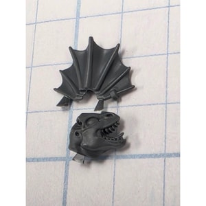 May include: A dark gray plastic miniature component consisting of a reptilian head with an open mouth showing teeth and a separate fan-shaped crest piece with ribbed detailing, both designed for assembly as part of a larger model figure.