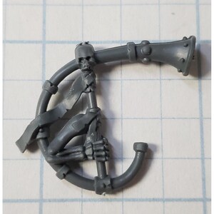 May include: A gray plastic miniature of a skeletal figure holding a horn. The horn is attached to a circular base. The figure is wearing a tattered cloth around its waist.