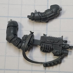 May include: A dark gray plastic miniature weapon and arm set, likely for a tabletop game. The set includes a detailed gun with an eagle emblem and a segmented arm piece. The pieces are unpainted and ready for customization.
