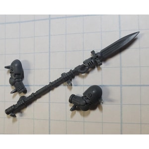 May include: A gray plastic spear with a pointed blade, detailed shaft, and two detached mechanical components. The spear is approximately 6 inches long. The components appear to be parts of a miniature model or figurine.