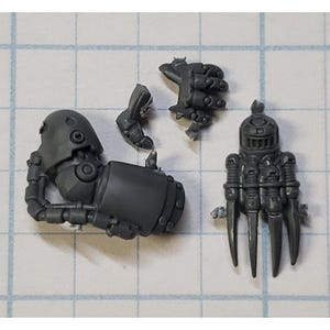 May include: A collection of gray plastic miniature parts, including a mechanical arm, a claw-like hand, and other components. The pieces appear to be from a model kit or a miniature figure, possibly for a tabletop game.