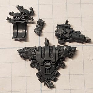 May include: A collection of unpainted, gray plastic miniature parts, including a skull and crossbones, a weapon, and a chest piece with wolf head details. These components are likely for a tabletop war game.