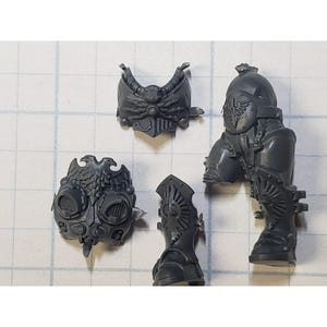 May include: A collection of unpainted, gray plastic miniature war game components. The pieces include a helmet, chest armor, a head, and a leg with a boot. The components feature detailed designs, including eagle emblems and ornate patterns.