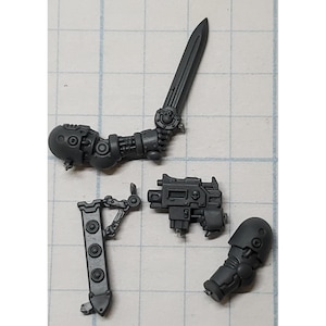 May include: A collection of gray plastic miniature parts, including a sword held by a mechanical arm, a pistol, a leg, and a piece with three circular details. The parts are likely for a model or miniature figure.