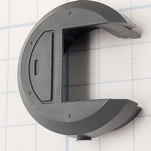 Puede incluir: Un componente de pl&aacute;stico gris semicircular con una puerta rectangular y una solapa con bisagras. El componente tiene varias peque&ntilde;as hendiduras circulares y est&aacute; sobre un fondo de cuadr&iacute;cula.