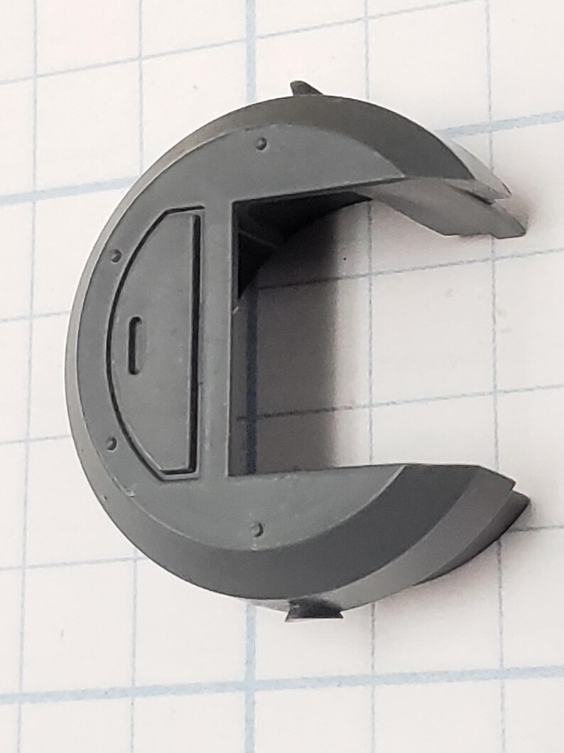 Puede incluir: Un componente de pl&aacute;stico gris semicircular con una puerta rectangular y una solapa con bisagras. El componente tiene varias peque&ntilde;as hendiduras circulares y est&aacute; sobre un fondo de cuadr&iacute;cula.