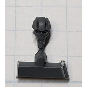 May include: A gray plastic miniature skull head with a raised design. The skull has a flat base and a slot for attaching to a model.