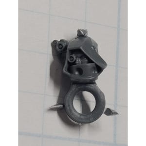 May include: A small, gray plastic component with a circular ring and a detailed top section. The top features a helmet-like shape with small, circular details and a textured surface. The ring has two small, pointed protrusions.