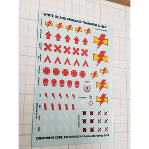 May include: A sheet of White Scars Primaris transfer decals. The sheet features red and yellow lightning bolts, arrows, skulls, and other symbols. Text includes "WHITE SCARS PRIMARIS TRANSFER SHEET" and "Games Workshop 2019".