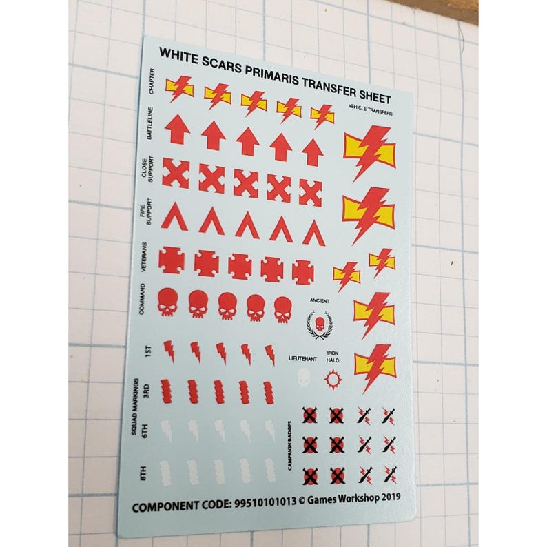 May include: A sheet of White Scars Primaris transfer decals. The sheet features red and yellow lightning bolts, arrows, skulls, and other symbols. Text includes "WHITE SCARS PRIMARIS TRANSFER SHEET" and "Games Workshop 2019".