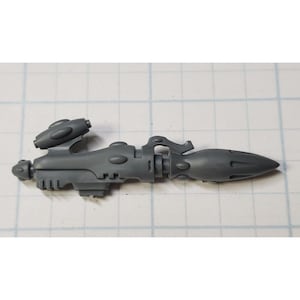 May include: A gray plastic model of a futuristic weapon with a barrel, a handle, and a scope. The weapon has a detailed design with various grooves and ridges.