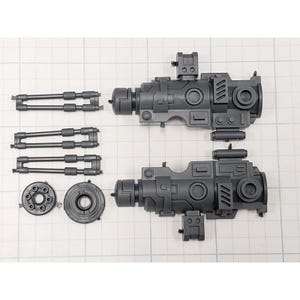 May include: Two gray plastic model gun components are displayed on a grid background. The components include two main gun bodies, two sets of barrels, and two circular pieces. The parts are designed for assembly.