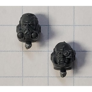 May include: Two gray plastic miniature heads. One head has a respirator mask, and the other has a single eyepiece. Both heads have a detailed, grim expression and are designed for miniature figures.