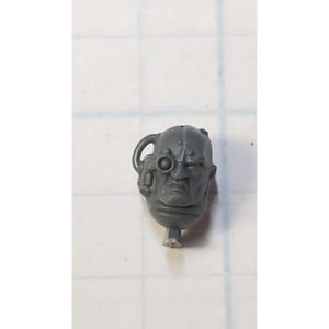 May include: A gray plastic miniature head with a single eye and a detailed face. The head is part of a wargaming set.