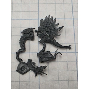 May include: A set of unpainted dark gray plastic miniature components including a reptilian creature with a large feathered headdress standing on a rock base, a separate reptilian leg piece, and a base section with a fern-like plant detail.