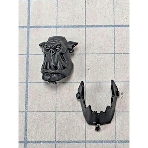 May include: A dark gray plastic miniature orc head component featuring prominent pointed ears, a flat nose, and a separate lower jaw piece with sharp teeth. The pieces are unpainted and molded in a matte finish, designed for assembly in tabletop gaming.