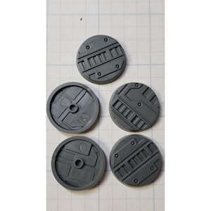 May include: Five gray plastic round bases with different geometric designs. The bases are for use in tabletop games.