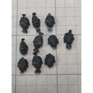 May include: A collection of ten dark gray plastic miniature heads, each featuring distinct facial expressions and molded details. These small gaming components are arranged in a grid-like pattern, showcasing various helmet designs and facial features.