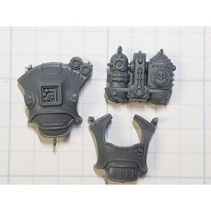 May include: Three gray plastic pieces for a model kit. The pieces are shaped like armor plates with intricate details and pipes.