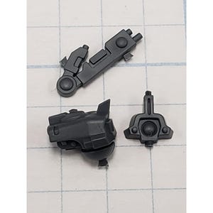 May include: Three dark gray plastic model kit parts are arranged on a grid-lined surface. The parts include a hinged arm, a rounded component, and a rectangular piece with a circular detail. These components appear to be for a model kit.