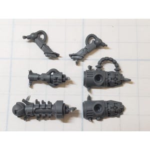 May include: A collection of six gray plastic miniature model parts, likely for a tabletop game or hobby project. The pieces include various mechanical components, such as tubes, cylinders, and what appear to be weapon attachments. The parts are arranged on a grid background.