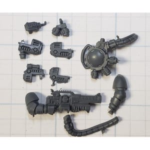 May include: A collection of gray plastic miniature model parts, including a large gun, various mechanical components, and a rounded piece with multiple attachments. The parts are arranged on a grid background.