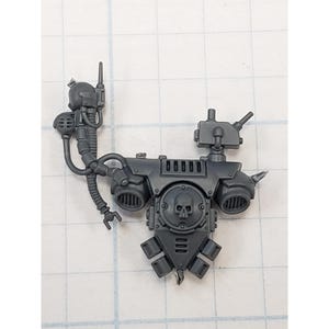 May include: A gray plastic miniature model component featuring a central panel with a skull emblem, flanked by two circular vents. It has various attached mechanical elements, including tubes, antennas, and a small robotic arm. The piece is designed for a tabletop game.