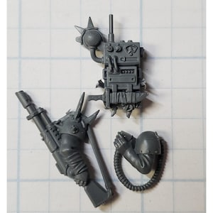 May include: A set of three unpainted gray plastic miniature components including a rectangular communications backpack with dials and an antenna, a long-barreled firearm with a spiked shoulder pad, and a bent arm with a connected flexible cable.