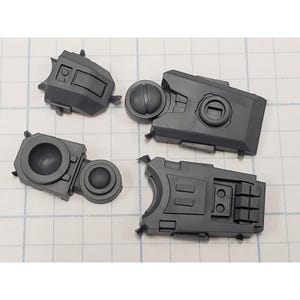May include: Four gray plastic model kit parts are arranged on a white grid surface. The parts have various geometric shapes, including rectangular and circular elements, with detailed surface features. The pieces appear to be components of a larger model.