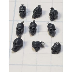 May include: Nine gray plastic skull figurines with a small detail on top of each skull. The skulls are arranged in a grid pattern on a white surface.