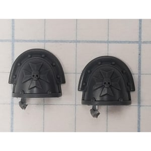 May include: Two dark gray shoulder pads with a skull and cross design. The shoulder pads have a curved top with rivets and a small attachment piece at the bottom. The design features a skull at the center of a cross.