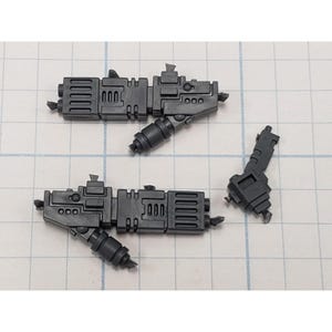 May include: Three dark gray plastic model gun parts are arranged on a grid. Two larger pieces have rectangular bodies with detailed sections and cylindrical attachments. A smaller piece is also visible.
