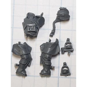 May include: Gray plastic miniature parts for a model kit. The parts include a torso, two legs, a head, and two small accessories.