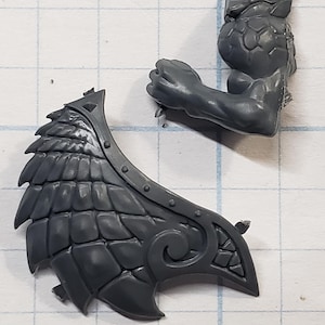 May include: Two unpainted plastic miniature parts: a detailed wing with a layered, textured design and a muscular arm with a clenched fist. The wing features an ornate, curved edge. The pieces are gray and appear to be for a model kit.