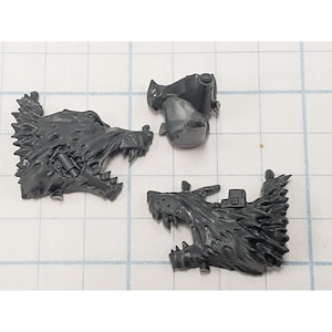 May include: Three unpainted dark gray plastic miniature components consisting of two stylized wolf head shoulder pads with open mouths and sharp teeth, and one small cylindrical connector piece with a rounded top and a flat base.