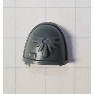 May include: Gray plastic miniature shoulder pad with a winged symbol and a small rectangular detail.