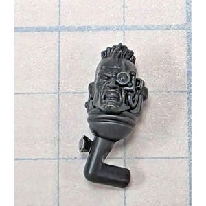 May include: A dark gray miniature plastic head featuring a mohawk hairstyle and a mechanical eyepiece over the left eye. The facial expression is intense with a bared-teeth grimace. The piece includes a neck joint and a protruding L-shaped connector.
