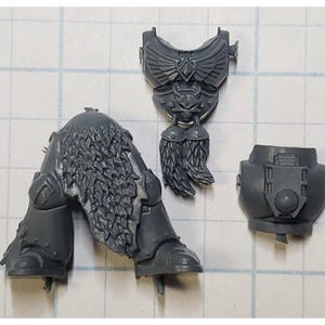 May include: A collection of unpainted, gray plastic miniature parts, including legs with fur detail, a chest piece with winged emblem, and a torso section. These components appear to be for a tabletop war game figure.