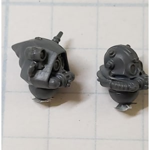 May include: Two gray plastic miniature helmet heads. One has a hood and multiple details, including a tube and two circular eye pieces. The other has a rounder shape with a breathing apparatus and similar details. Both are on a white surface.