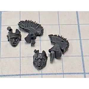 May include: Four unpainted dark gray plastic miniature components consisting of two distinct head pieces and two separate crests. One head features a human-like face, while the other displays a mechanical or armored mask with a single visible eye lens.