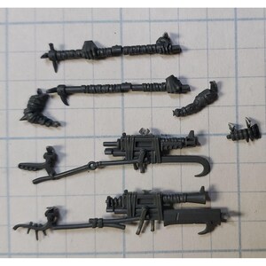May include: A collection of gray plastic miniature weapon parts, including spears, hooks, and gun-like components. The pieces are arranged on a grid-lined surface, suggesting a model-making or hobbyist context.