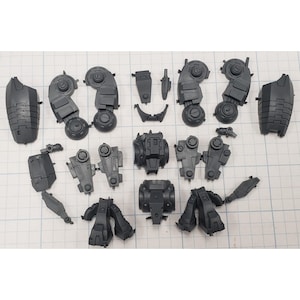 May include: A collection of unassembled dark gray plastic model kit parts, including various mechanical components such as leg armor, joints, and body sections. The parts are arranged on a grid-lined surface.