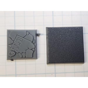 May include: Two gray plastic square bases for miniature wargaming. One base has a raised design of cracked pavement with the word "Dead" printed on it. The other base is plain.